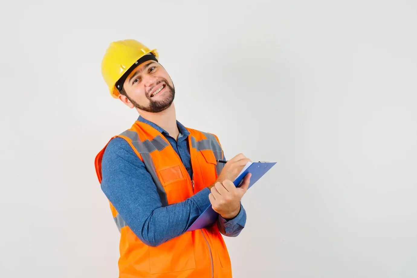 Front view of a young builder in a shirt, vest, and helmet, taking notes on a clipboard and looking cheerful.